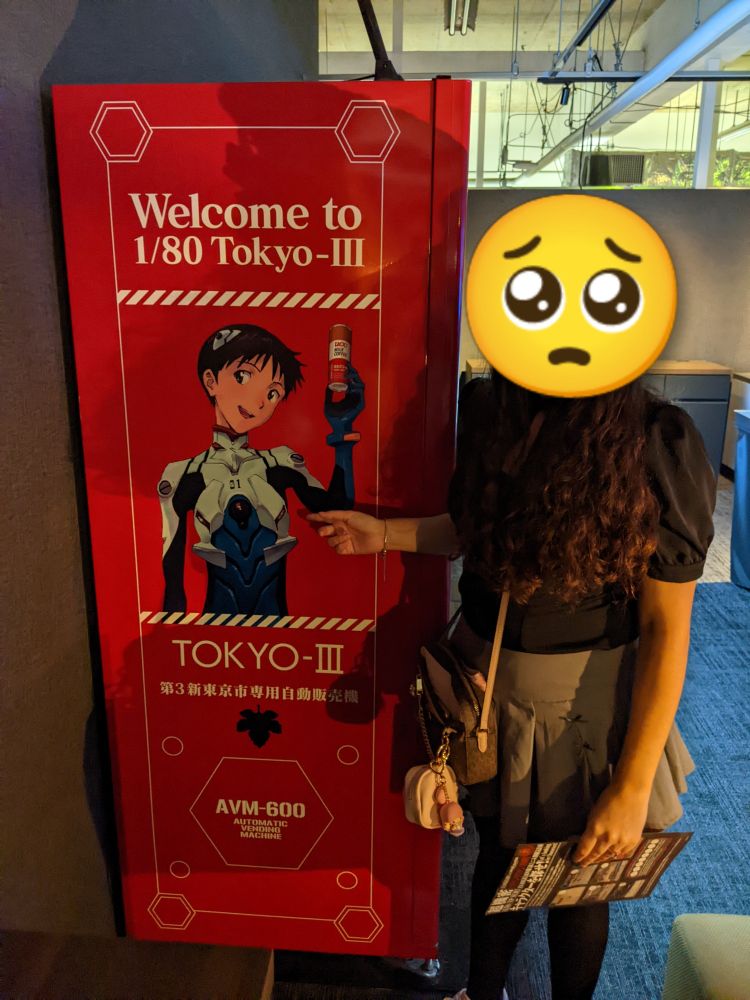 @meaVT standing next to a vending machine with shinji ikari from evangelion on it and pointing at him awkwardly. Her face is censored by a pleading emoji