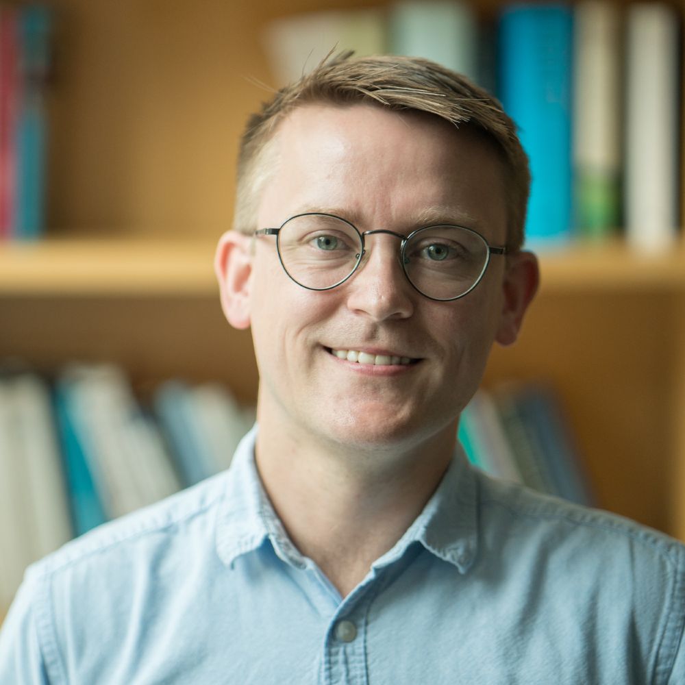 David Lydon-Staley, wearing round glasses and a light blue button-up shirt, smiling in front of a wooden bookshelf filled with books