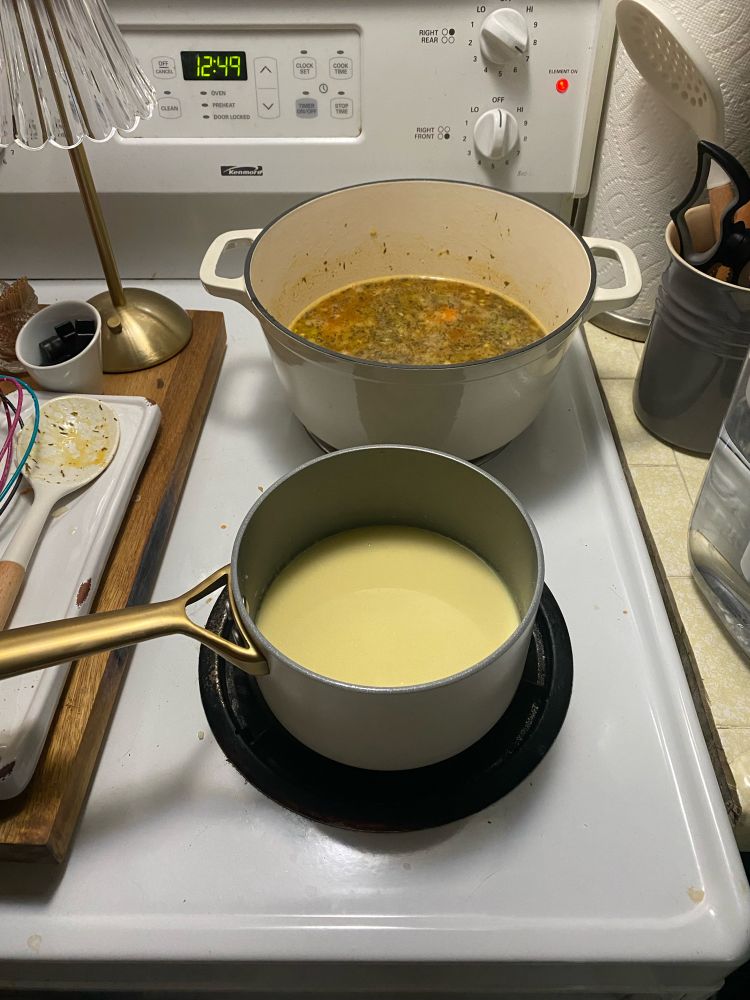 The mirepoix has become soup base in the Dutch oven on a back burner while milk in a roux thickens on the front burner. Dirty spoons and a whisk are off to the left side 