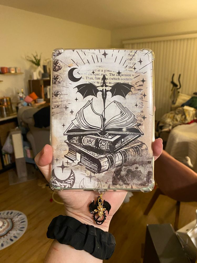 A kindle is being held up with a cover insert showing two dragons over a stack of books, representing The Empyrean series by Rebecca Yarros. A kindle charm with a black dragon and a gold dragon dangle from the kindle’s charging port 