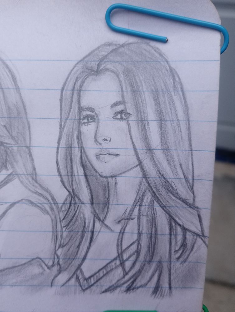 A pencil sketch of a young woman with long, flowing hair and a calm, confident expression. She is looking slightly to the side, with detailed shading on her hair and face. The drawing is done on lined notebook paper, held in place with a blue paperclip at the top right corner.