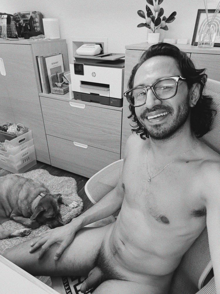 A naked person sitting in an office chair at their desk smiling at the camera. A dog is laying next to them on a dog bed with bookshelves behind them. The picture is black and white. 