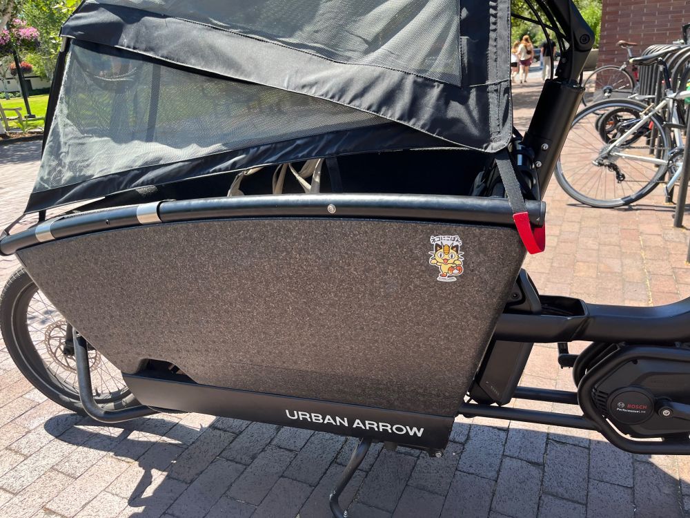 An urban arrow cargo bike with a sun cover and Meowth Pokémon sticker.