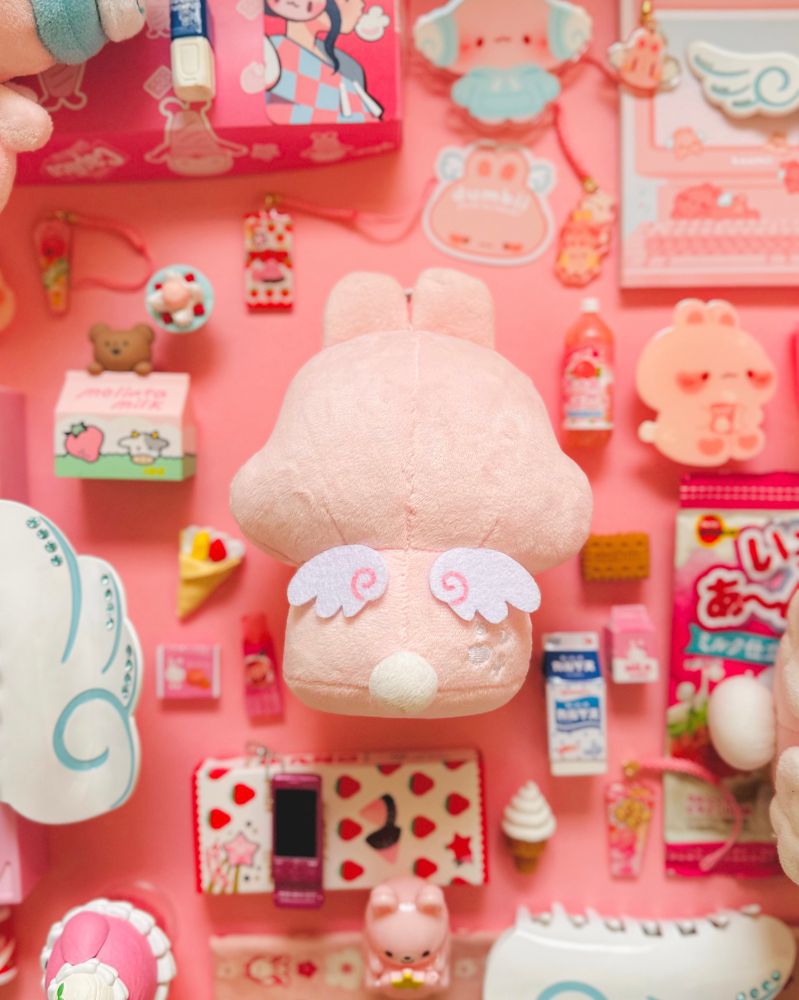 digital flatlay photo of a soft light pink rabbit keychain plush’s back view. the bun has a pair of angel wings with pink swirls inside, a small white pompom tail and white sparkle embroidery on its right side. surrounding the plush are knickknacks such as little toy trinkets, food shaped erasers, hair claws and phone charms. the background color is medium blush pink. 

the plush is for preorder at baomii.com
early bird discount ends 2/28
