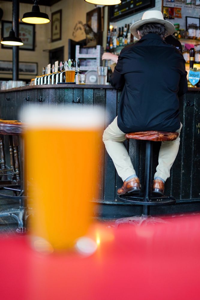 Blurry pint in foreground with a well dressed man sat at the bar in the background