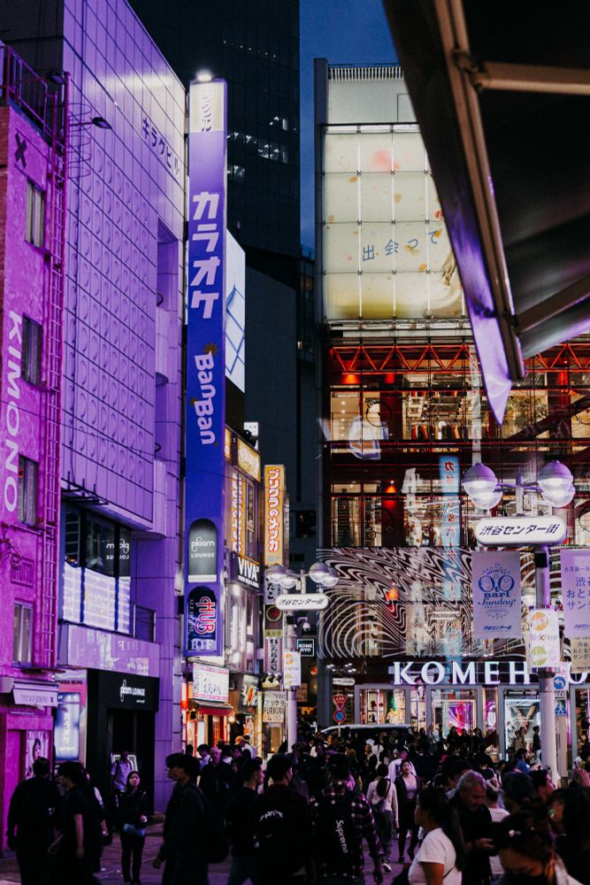 Photo of a busy main street in Tokyo either hundreds of people walking around. There are tons of neon signs on basically all walls.