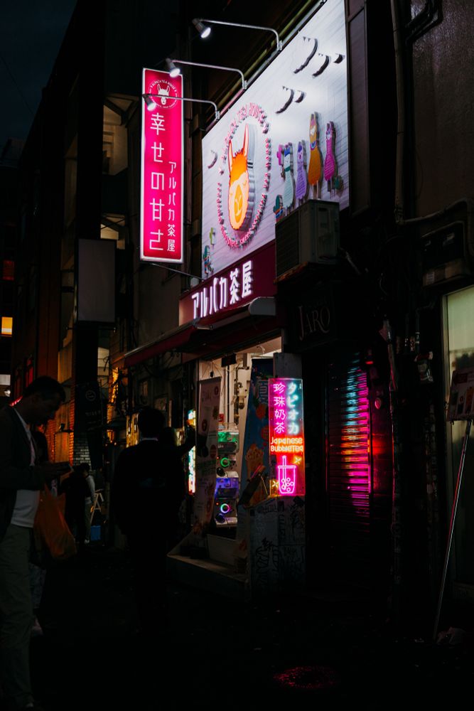 Photo of a small side street at night with some people hanging around. One shop has bright neon signs.