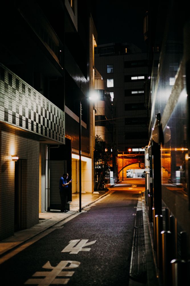photo of a small, narrow side street of Tokyo, barely lit, with a person standing holding a phone