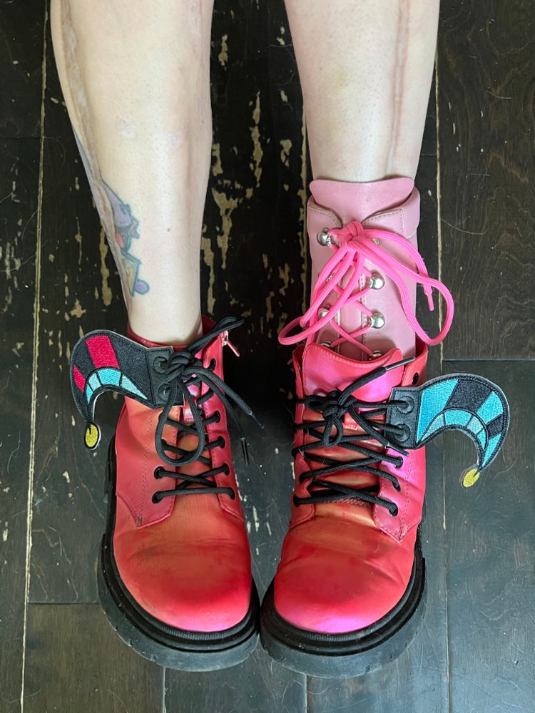 Photo showing person with a light pink prosthetic wearing hot pink shoes.  On the oute sides of their shoes are shoe wings designed to look like the jester’s cap Helluva Boss’s Fizzarolli wears.