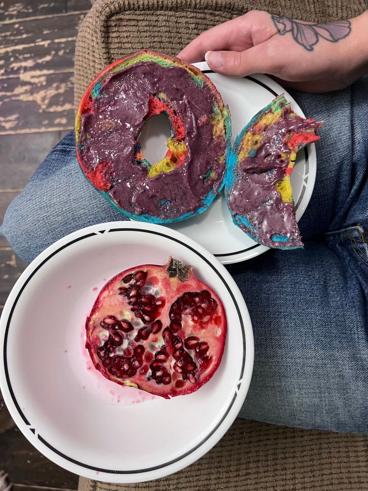 Bowl containing half a pomegranate and a plate with a rainbow bagel with purple creamed honey spread 
