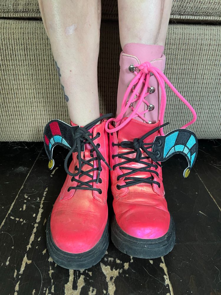Photo showing person with a light pink prosthetic wearing hot pink shoes.  On the oute sides of their shoes are shoe wings designed to look like the jester’s cap Helluva Boss’s Fizzarolli wears.