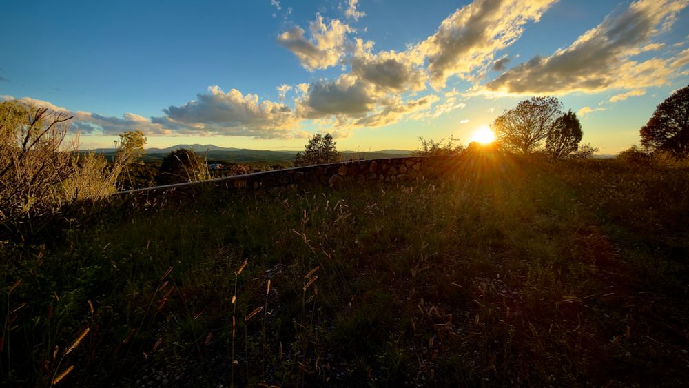 Clouds in a blue sky surrounding the sun setting on the horizon above distant violet colored mountains illuminating long yellow strands of grass against the dark earth in the foreground.