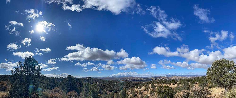Panoramic image of bright blue skies with puffy white clouds and a bright sun above a landscape with trees and mountains in southwestern New Mexico near Silver City.