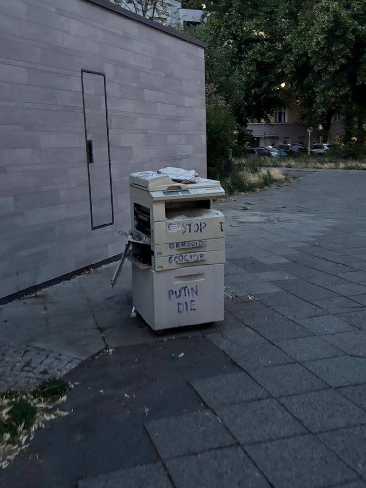 Büro-Kopierer vor einer öffentlichen Toilette stehend, auf einem Bürgersteig in Berlin Neukölln mit verschiedenen Parolen bemalt.