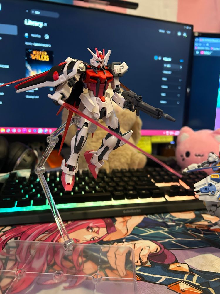 completed gundam model kit on an aerial stand to simulate flight poses. the mecha is primarily white, with red and pink accent pieces on the torso, head, and feet.