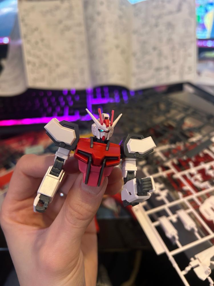 a hand holding a partially constructed white, pink, and red gundam model. only the torso, head, and arms have been built.