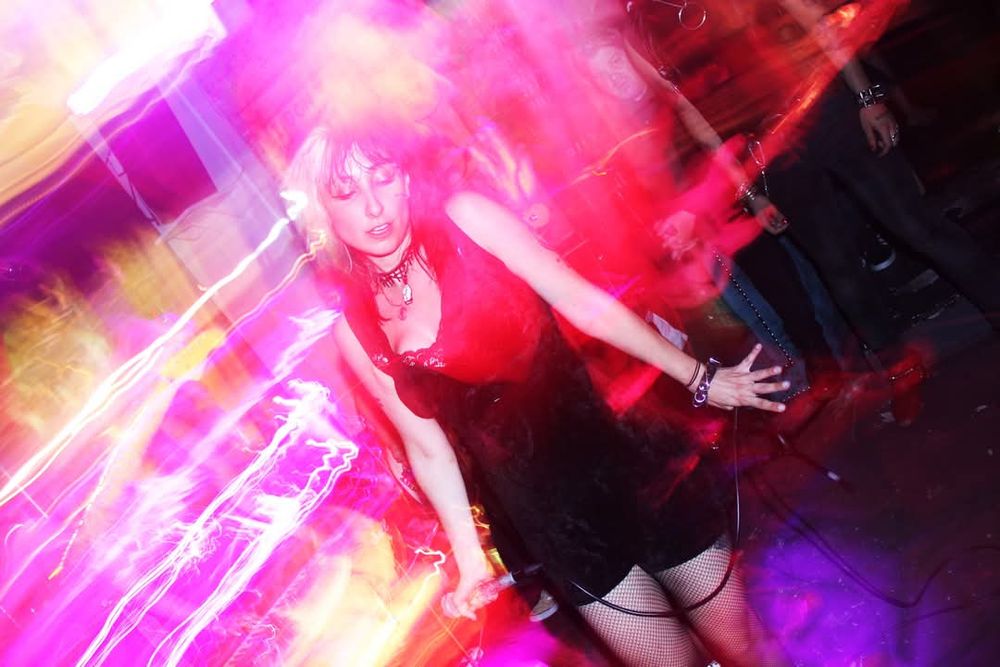 Long exposure of Diana from Dame. There are pink, yellow, and red streaks of light behind her. Diana has black hair with a blonde streaks, and it wearing a sleeveless black outfit, and holding a mic.