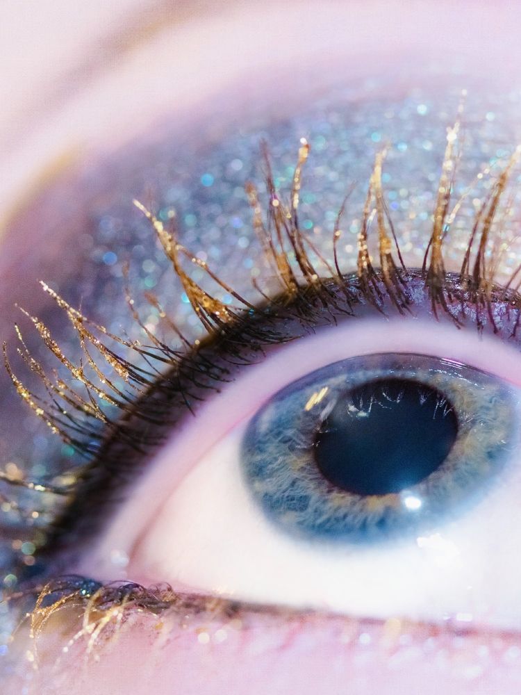 A close-up of a left eye. The iris is mostly blue with flecks of greenish yellow towards the center. The top and bottom lashes start black but have a glittering gold color on the tips. The upper eyelid is has a shimmery blue eyeshadow.