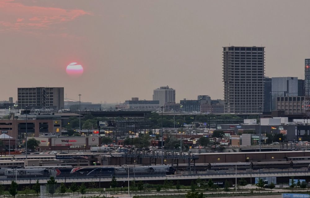 Red sun near city horizon partly obstructed by tendril of cloud. Grey sky.