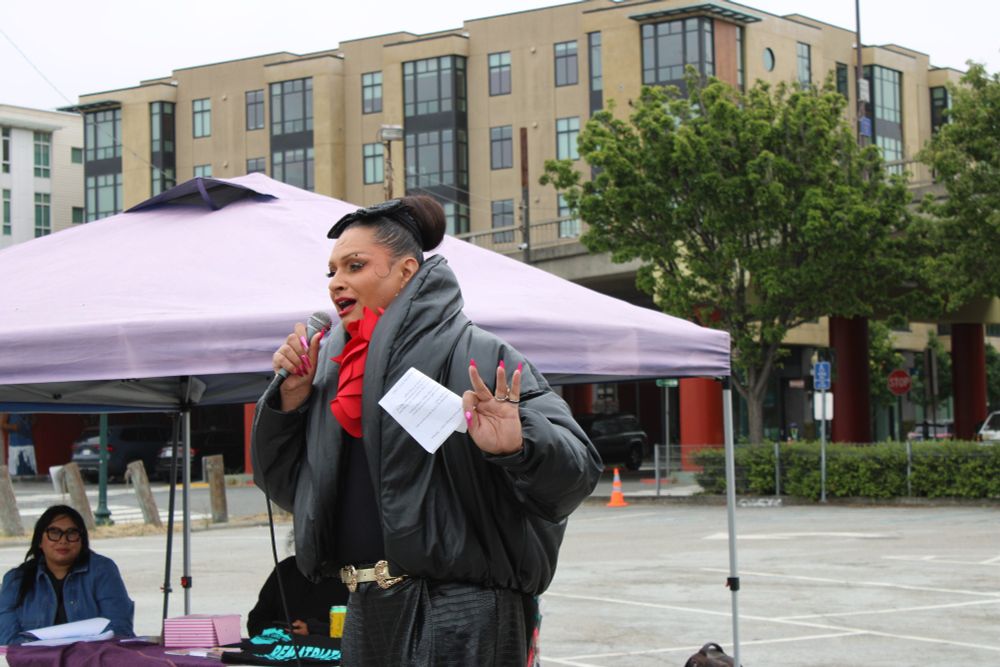 Per Sia is dressed in black and red with red finger nails and a top bun. In the background a tent with staff of the Sogorea Te Land Trust.