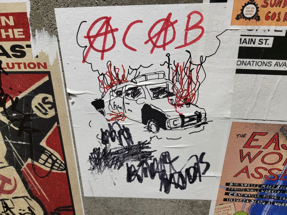 ACAB poster with burning cop car and circle As on concrete street post with other posters.