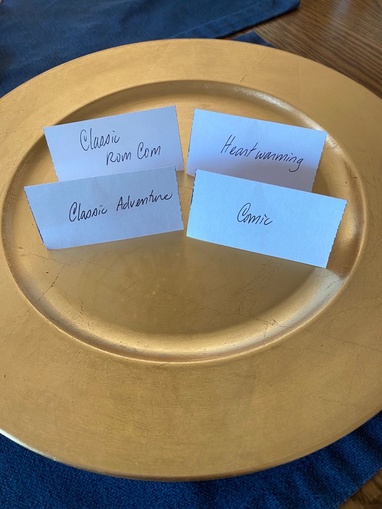 A gold plate with 4 folded cards on it that read “classic rom com”, “heartwarming”, “classic adventure” and “comic”