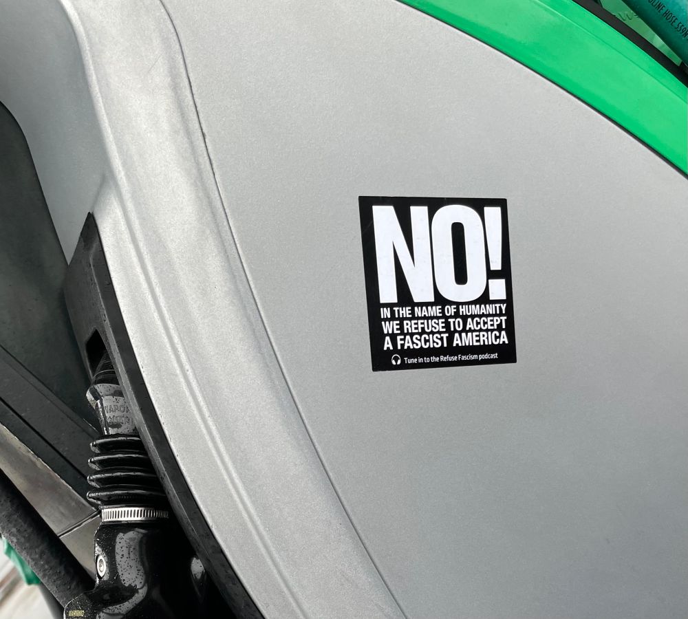 Photo of the side of a gas pump with a sticker on it saying “NO! In the name of humanity we refuse to accept a fascist America” in white text on black background 