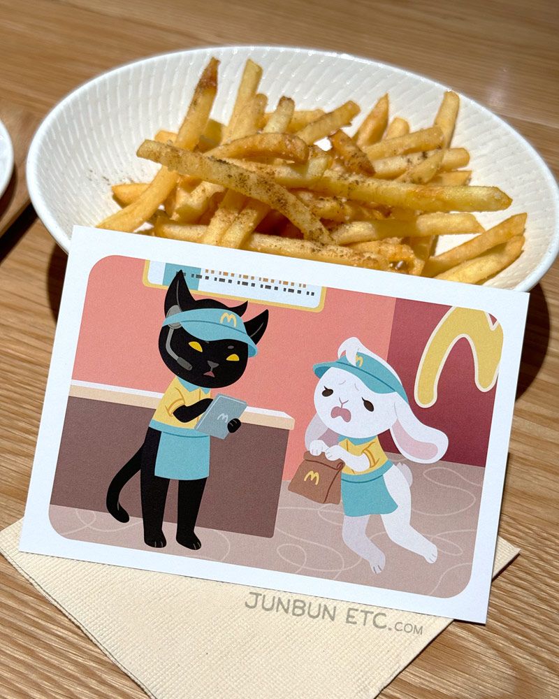The photo shows the MewBunny's postcard image depicting JunBun - the white rabbit mascot - and MaxCat - the black cat mascot - as fast food/diner/burger joint minimum wage workers, complete in MewBunny's uniforms and visors. MaxCat is reviewing orders on a tablet, and JunBun is falling over in dismay with a brown paper bag in paw. Behind is a plate of soy-sansho fries. 