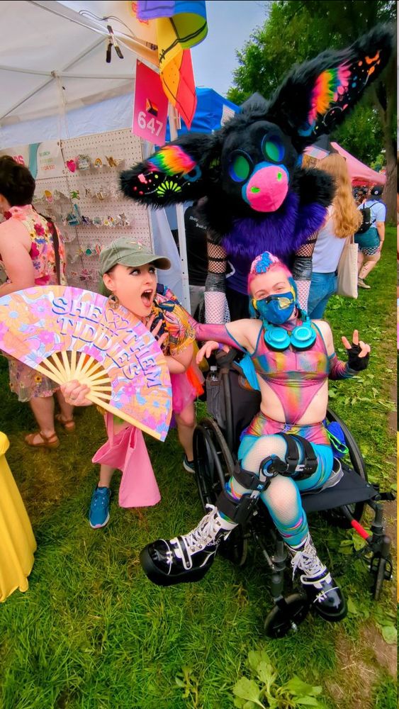 an image of a person in a bright outfit in a wheelchair with a fursuiter behind while a person with a colorful fan saying She Them Tiddies stands to the side.
