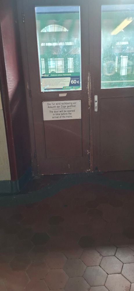 Braune Tür ohne Griff mit zwei Glasfenstern zwischen Wartehalle und schmalem Bahnsteig, darunter ein Schild mit Text in Deutsch und Englisch zur Öffnung der Tür vor Zugankunft: 'Die Tür wird rechtzeitig vor Ankunft der Züge geöffnet. The door will be opened in time before the arrival of the trains.' 