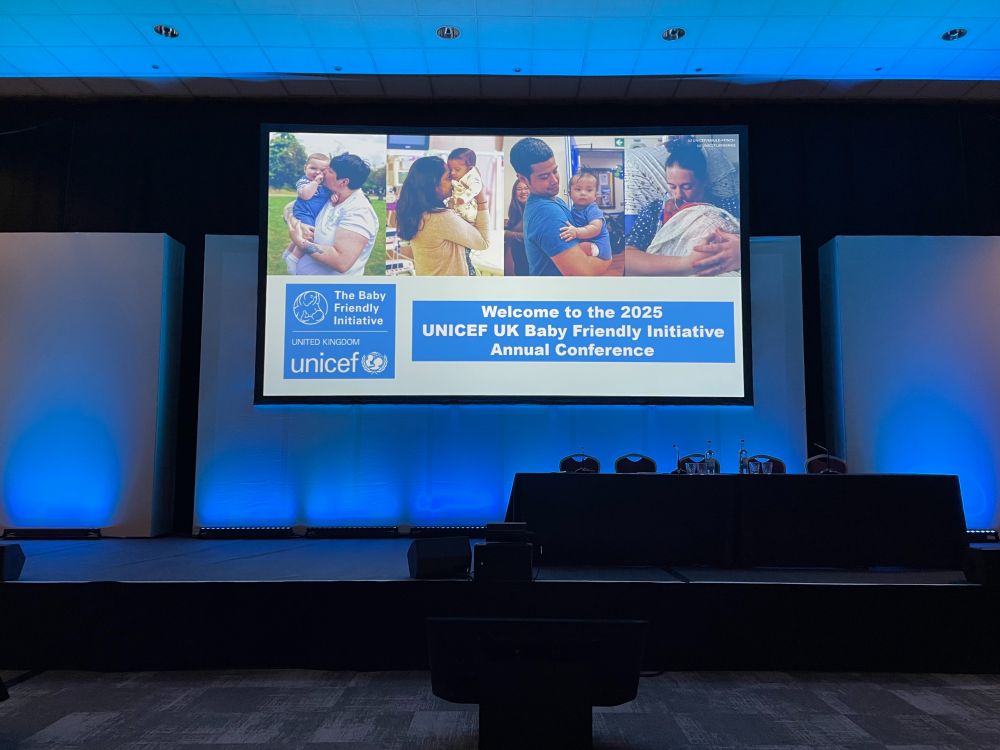 The stage at the conference centre ready to start the day with a welcome slide saying Welcome to the 2025 UNICEF UK Baby Friendly Initiative Annual Conference