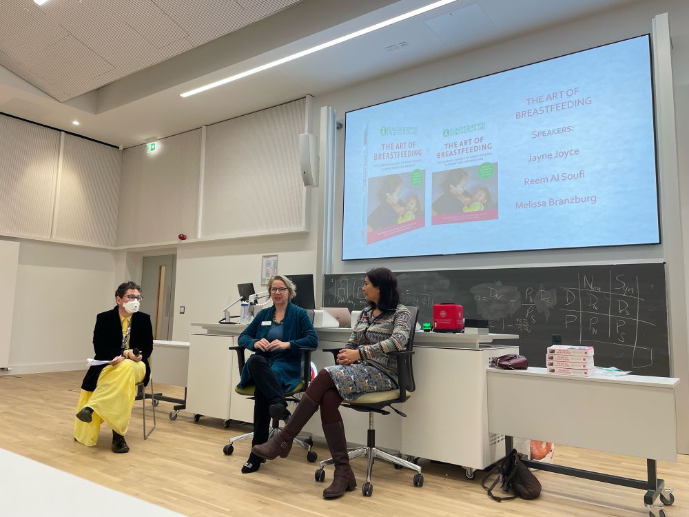 Melissa Branzburg, Jayne Joyce and Reem Al Soufi sitting at the front of a lecture hall at the University of Edinburgh with a slide in the background showing the UK cover of The Art of Breastfeeding 