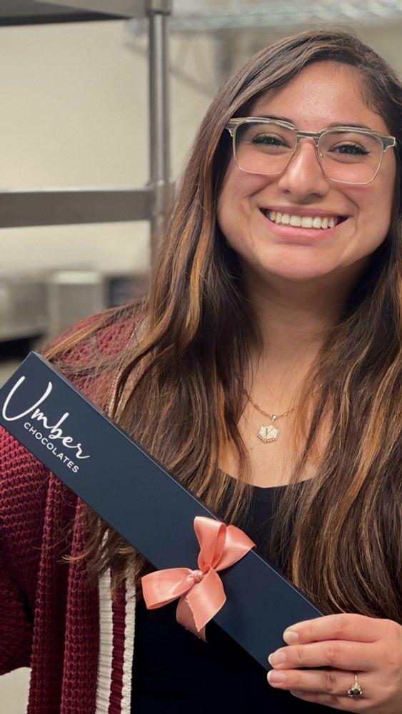 Owner of Umber Chocolates, Veronica, smiling while holding a 9 piece box of chocolates with a bow. 