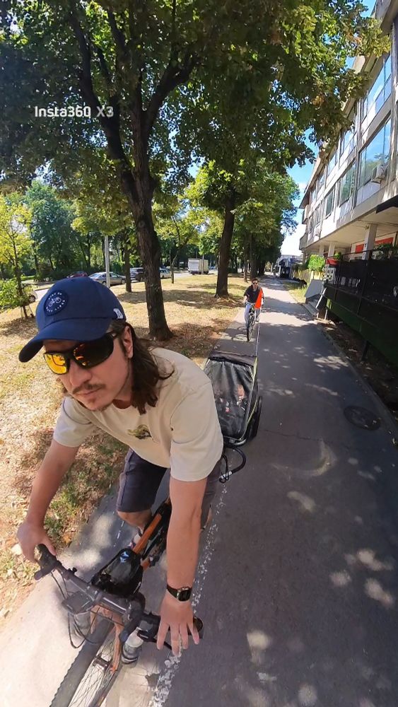 Father on the bicycle towing two small babies in a trailer and mother following them on her bike.