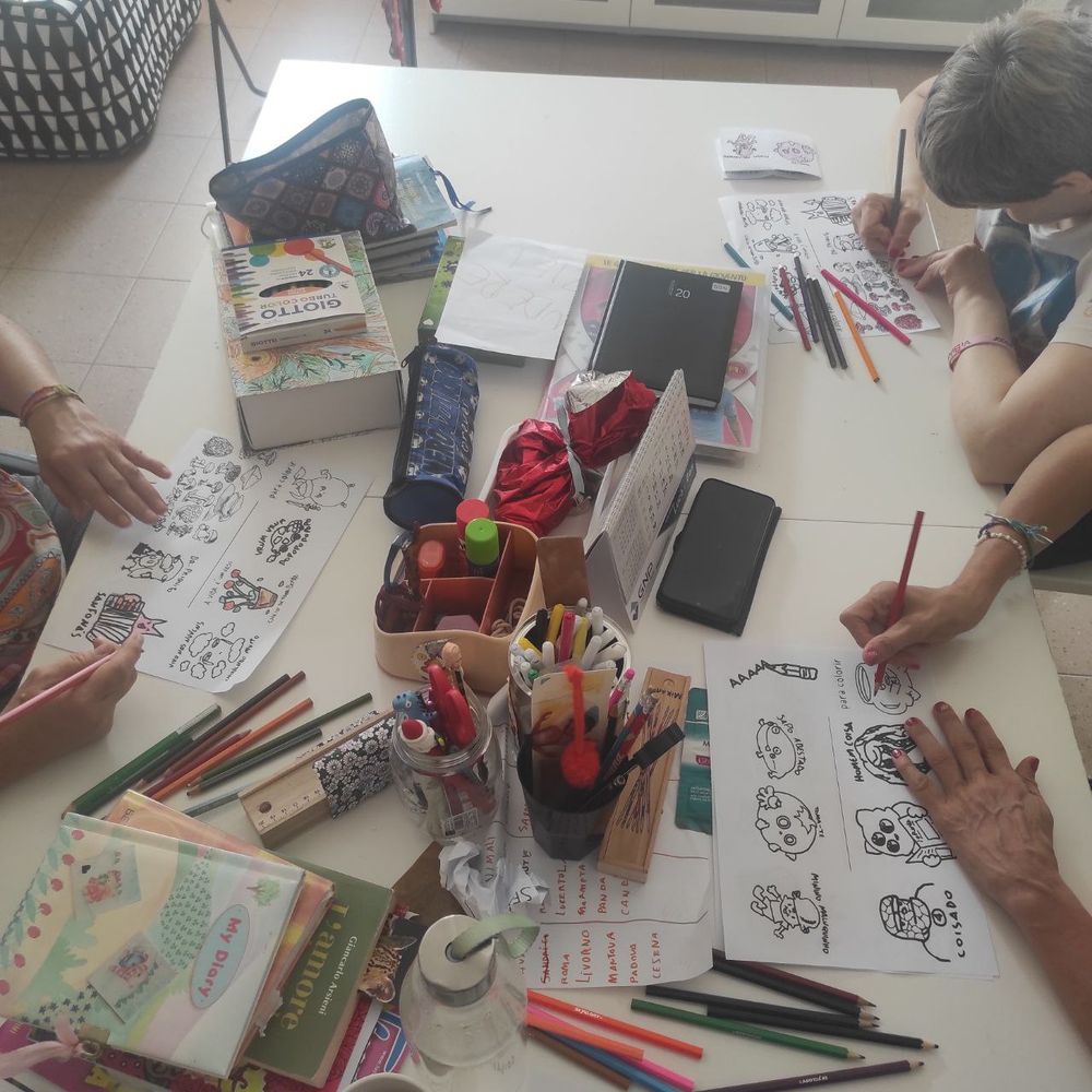 Foto de uma mesa, cheia de material de papelaria, com pessoas pintando desenhos