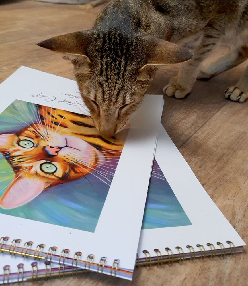 an oriental cat inspecting some 2026 Painted Cat calendars. The calendars have a painting of a bengal cat on the front.
