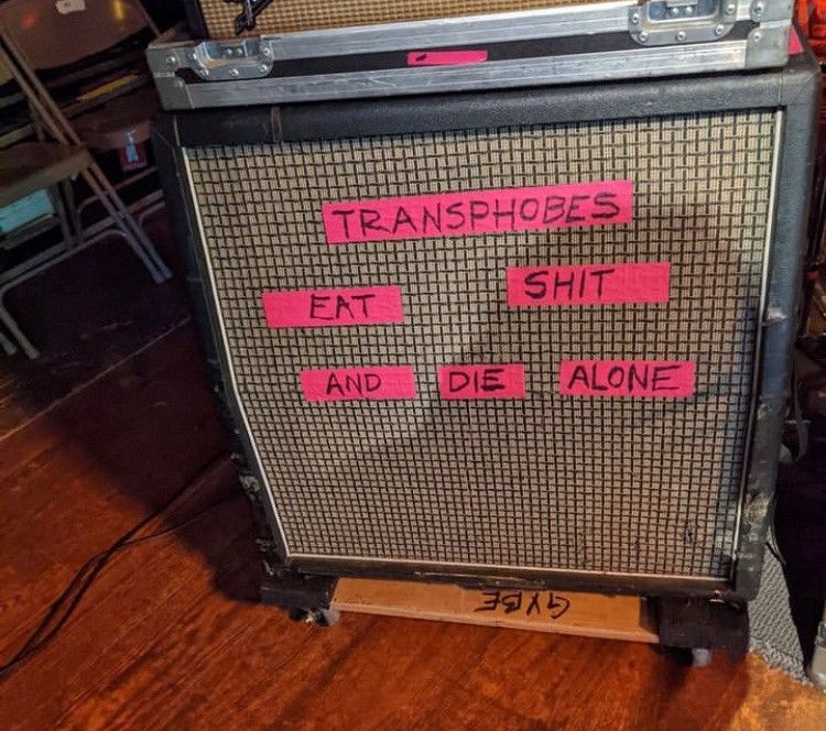 A guitar speaker cab with pink tape stuck across it. The words "transphobes eat shit and die alone" are written on the tape.