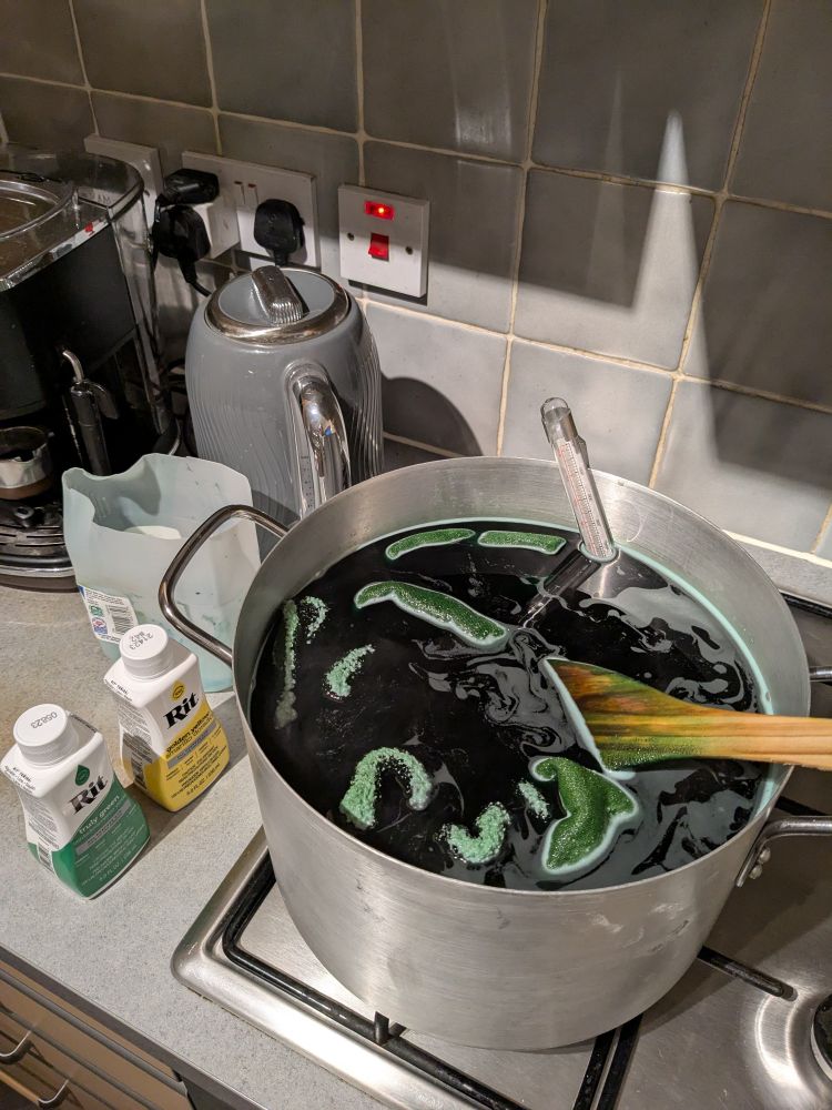 A large pot on a gas stove. It's full of dark green liquid with a few folds of cloth breaking the surface, and a green-stained wooden spatula. There are bottles of yellow and green fabric dye on the worktop next to it. 