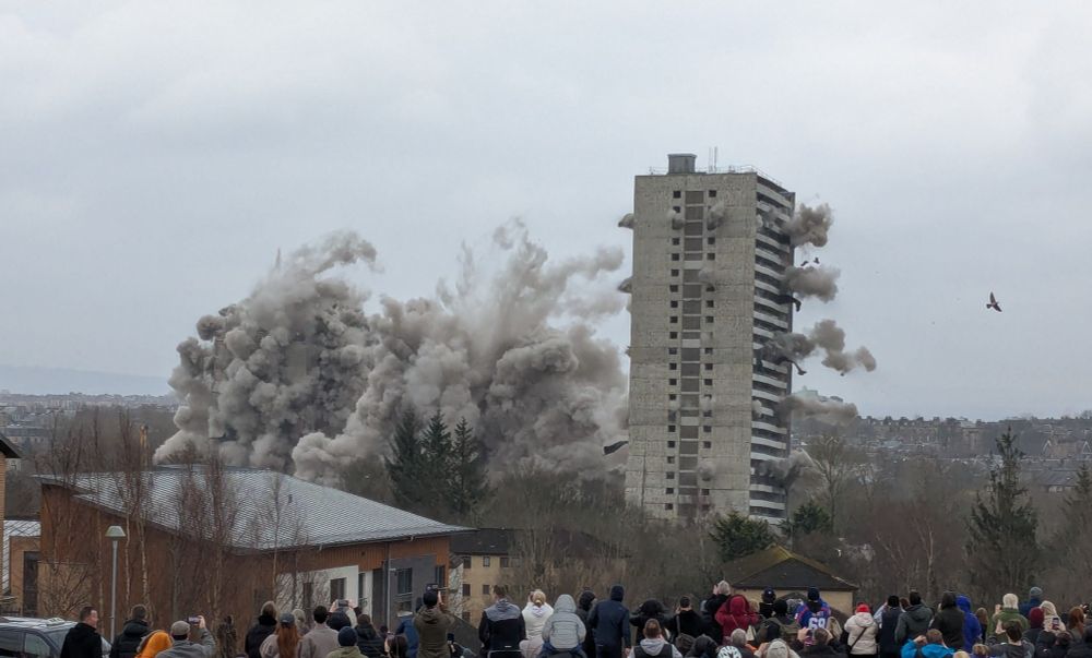 Three tower blocks being demolished on an overcast day. The one on the right is just beginning to fall, while all that's visible of the other two is plumes of grey dust.