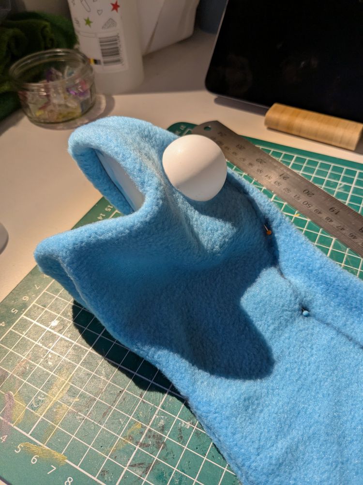 The head of a Kermit-style frog puppet, made of blue fleece (shout out to the Mandors remnant bin!). It's lying on my desk full of pins and sharpie lines while I figure out how to get the shape and size right. 