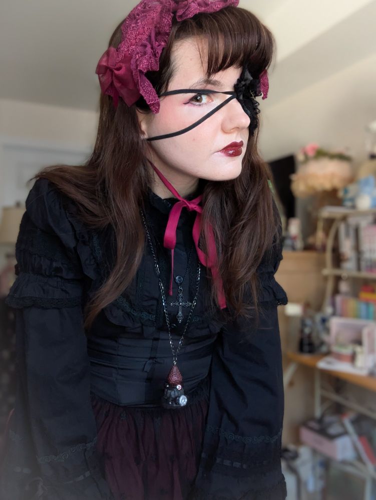 A woman with brown hair and fringe wearing a black and bordeaux gothic lolita coord and an eye patch. She is making a neutral expression at the camera.
