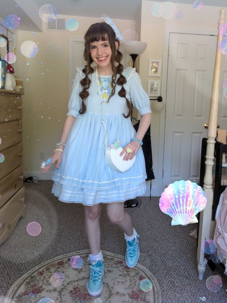 A brunette woman with bangs and long brown hair wearing bubble pigtails. She is smiling while wearing a simple sax blue lolita coord.