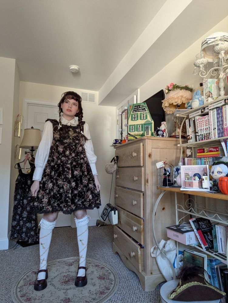 A woman in an old school lolita coord. She has on a brown, floral corduroy jsk, an off white blouse with long sleeves and a peter pan collar, an oversized heart locket necklace, knee length socks with lace on top and Mary Jane shoes with a heel. She also has an off white headdress with butterflies.
