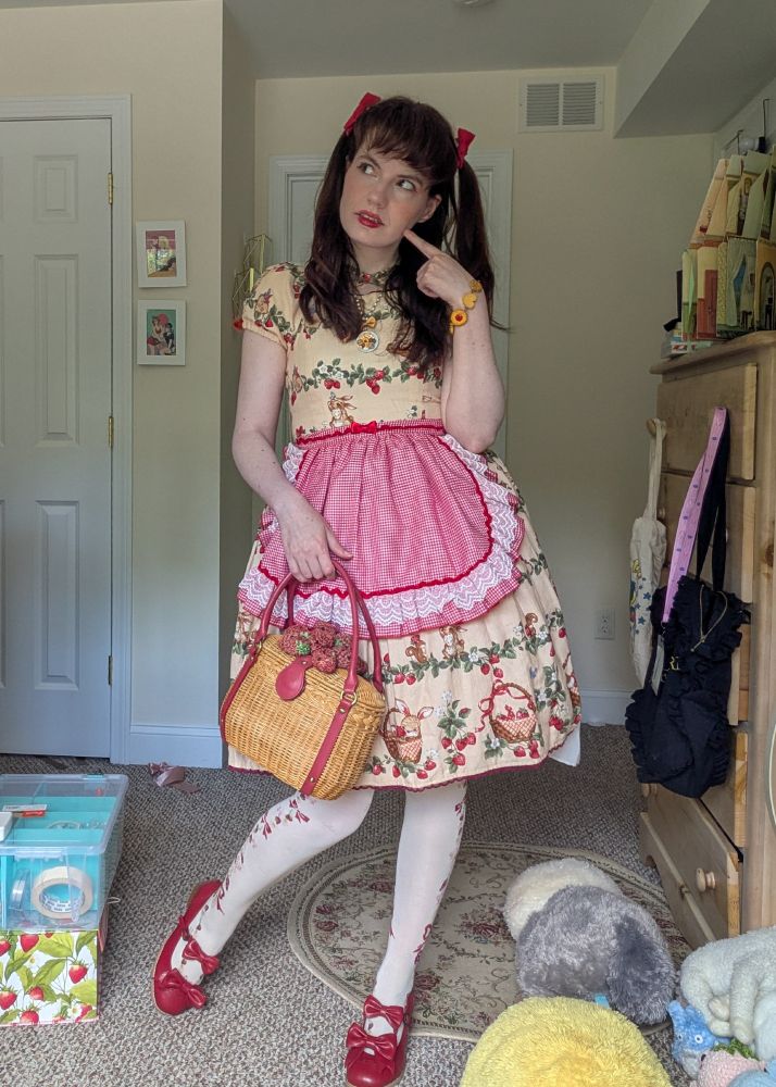 A brunette woman in a classic-sweet lolita coord. There's two mini pigtails in her hair with red ribbon securing them and she's wearing an ivory, knee length dress with a print of small forest animals, strawberries and baskets. She has a red gingham rounded apron with lace and is carrying a wicker basket with strawberries on it. Her tights are a lighter ivory with a print of red and pink strawberries as well as bows. She has red shoes on. She has a small necklace with sweets on it and a matching bracelet.