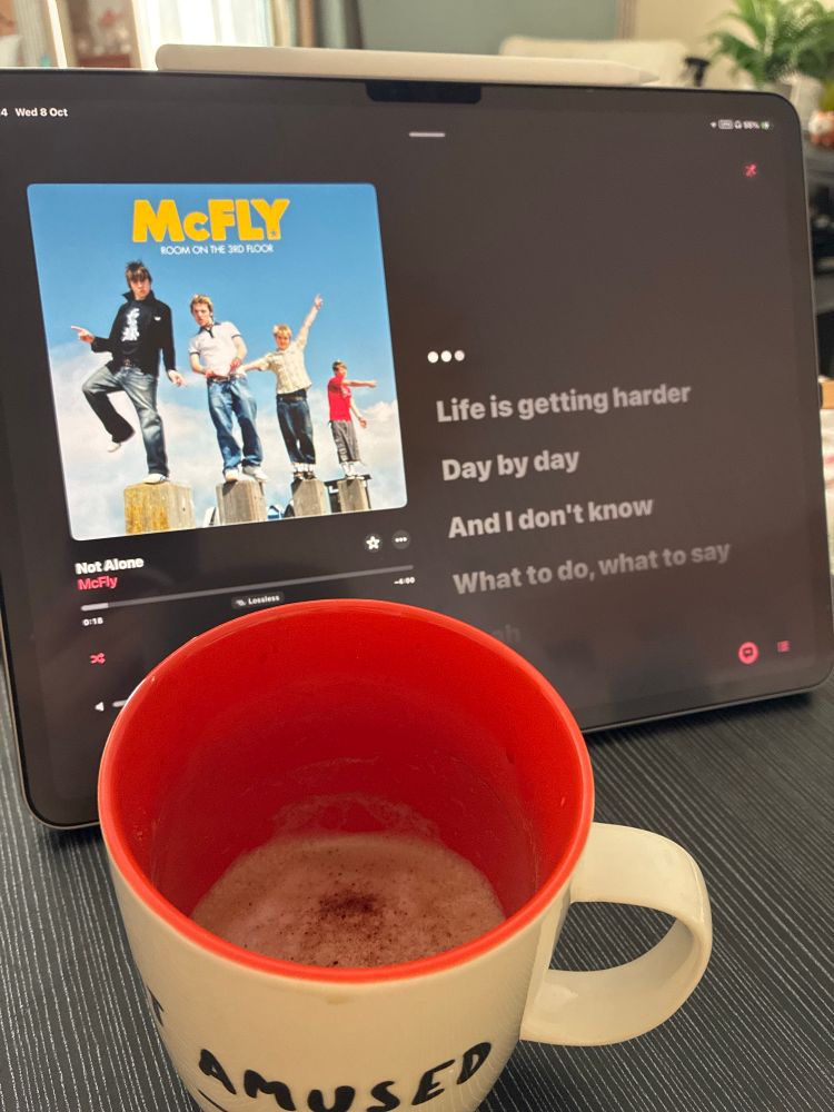 A photo of white cup with a red interior on a black wood effect tabletop, looking down from a high angle. The cup almost empty, a cappuccino with chocolate power sprinkled on top of foamy milk. Behind stands an iPad showing a music player and lyrics. The song playing on the screen is Not Alone by McFly 