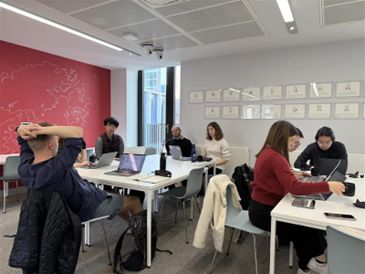 A classroom, in which several people are sat around two desks with laptops in front of them. Some are looking at their laptop screen and others are looking at each other, as though they are discussing something. 