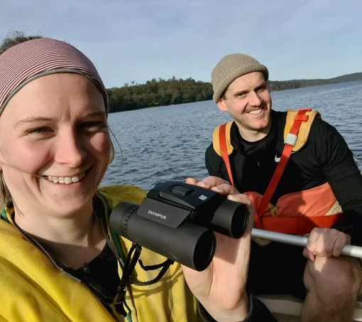 📷 Kelsey Picard - taken during Where? Where? Wedgie! surveys. A man and a woman are sitting in a double kayak on a bright day in open water. They are wearing life jackets and smiling at the camera. The man, behind, holds a paddle, while the woman, in front, holds the camera for the selfie and a pair of binoculars in the other hand.