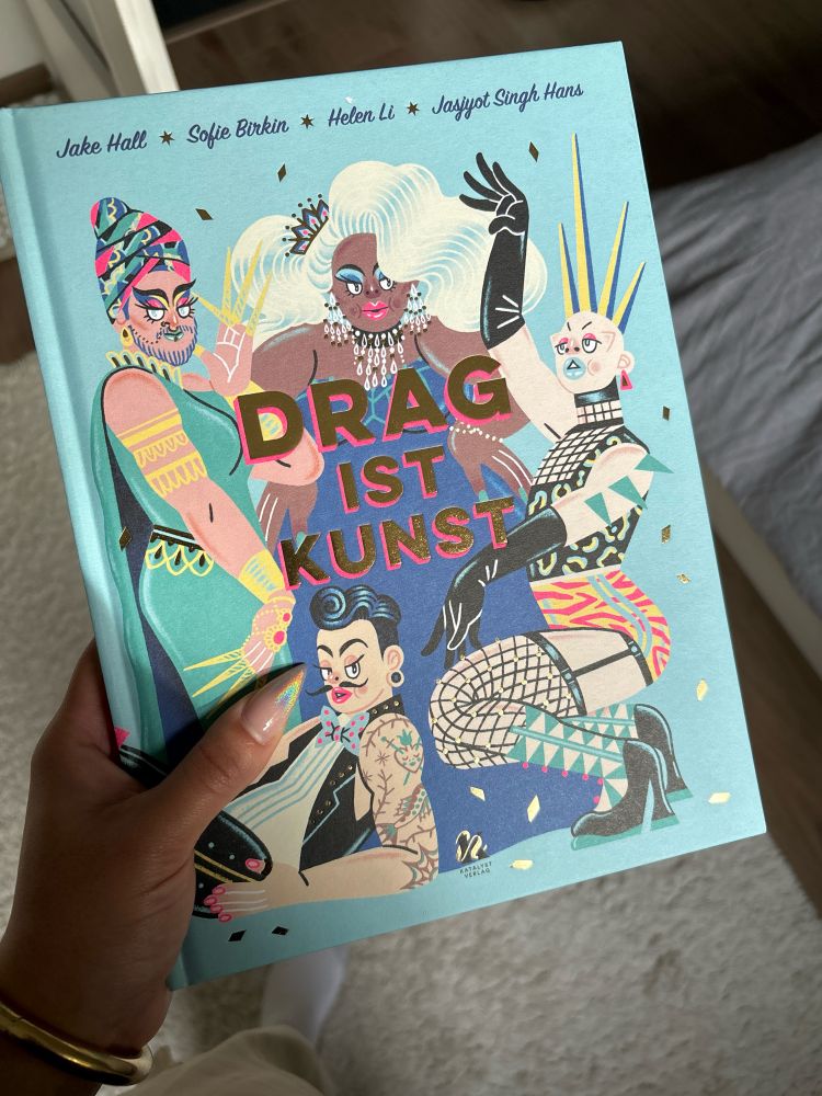 Buch mit dem Titel „Drag ist Kunst“ von Jake Hall et al. Auf dem Cover ist der Titel zentral in goldenen Lettern aufgedruckt, dahinter sind kündtlerische Abbildungen vier verschiedener Drag Queens extravaganten Outfits abgebildet.