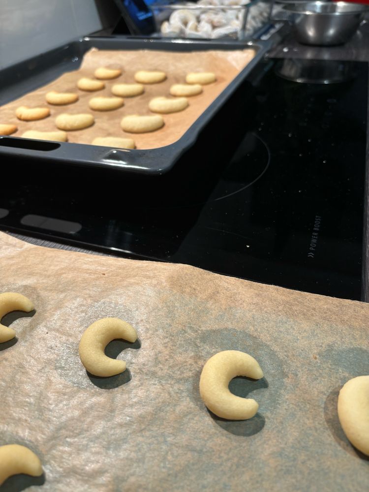 Vanillekipferl in 3 Stufen: im Vordergrund frisch geformte Teighörnchen, mittig auf dem einem Blech frisch gebacken aus dem Ofen und unscharf im Hintergrund in Puderzucker gewälzt.