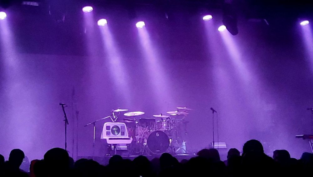 Bühne in violettes Licht gehüllt. Publikum im Vordergrund.